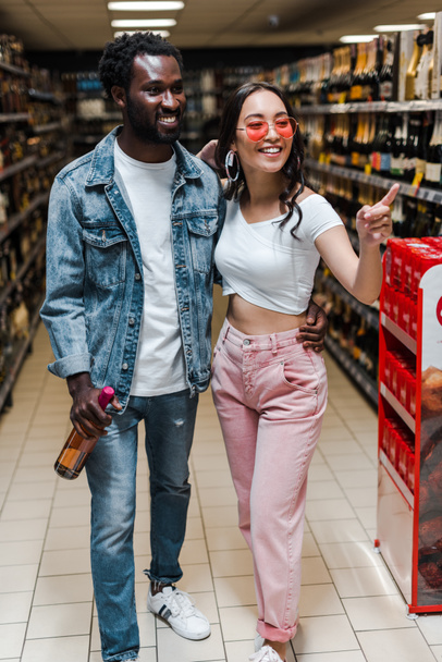 šťastný afroamerický muž držící láhev poblíž asijské dívky v paprskových brýlích ukazujícími prstem v supermarketu  - Fotografie, Obrázek