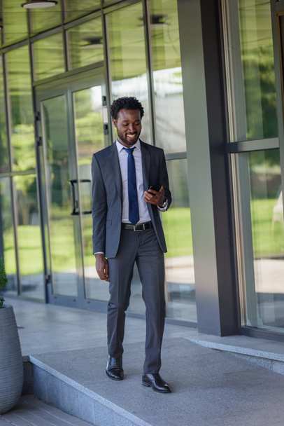 オフィスのブイディングに沿って歩いている間、スマートフォンを持つ笑顔のアフリカ系アメリカ人ビジネスマン - 写真・画像