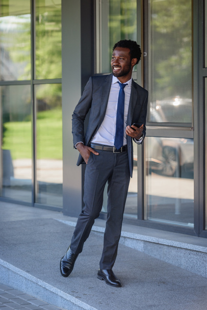 cheerful african american businessman with smartphone walking along office building and holding hand in pocket - Foto, Imagen