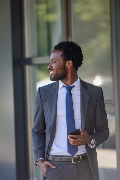 sonriente hombre de negocios afroamericano con teléfono inteligente cogido de la mano en el bolsillo y mirando hacia otro lado
 - Foto, Imagen