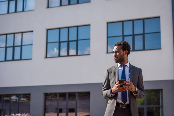 スマートフォンを持ちながら目をそらす自信のあるアフリカ系アメリカ人ビジネスマン - 写真・画像
