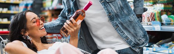 panoramic shot of happy asian girl in sunglasses holding bottle with wine while sitting in shopping cart near african american man  - Foto, Imagen