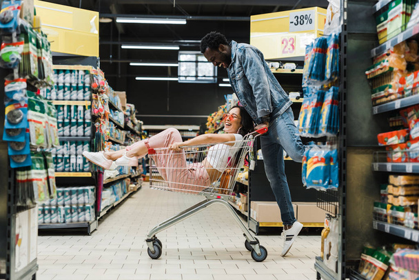 glücklich afrikanisch-amerikanischer Mann schaut asiatisches Mädchen an, das im Einkaufswagen im Supermarkt sitzt  - Foto, Bild