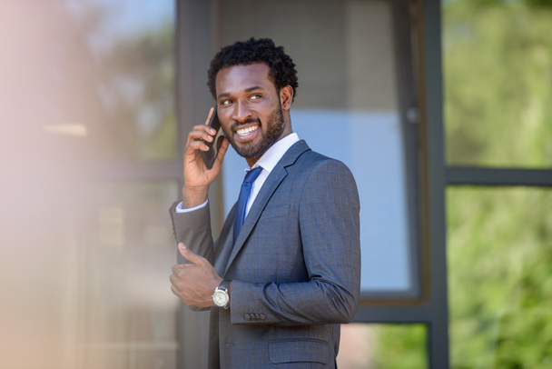 Selektiver Fokus eines lächelnden afrikanisch-amerikanischen Geschäftsmannes, der in der Nähe eines Bürogebäudes mit dem Smartphone spricht - Foto, Bild