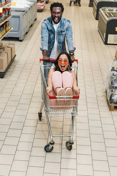 Gelukkig African American man wandelen met Aziatische meisje in zonnebril zitten in winkelwagen  - Foto, afbeelding