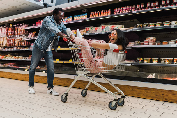 excited african american man near happy asian girl in sunglasses sitting in shopping cart  - Photo, Image