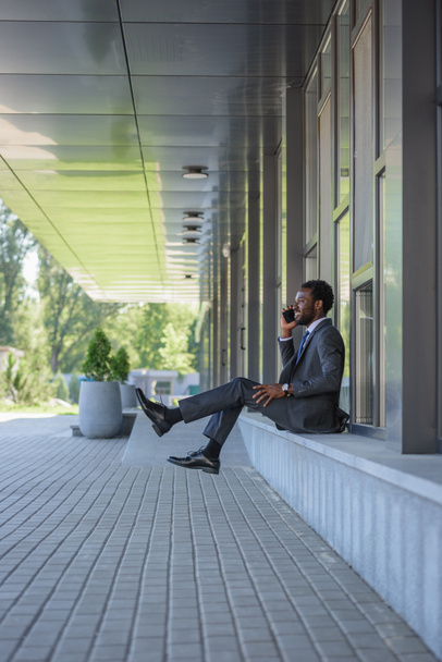 alegre hombre de negocios afroamericano hablando en el teléfono inteligente mientras está sentado cerca del edificio de oficinas
 - Foto, imagen