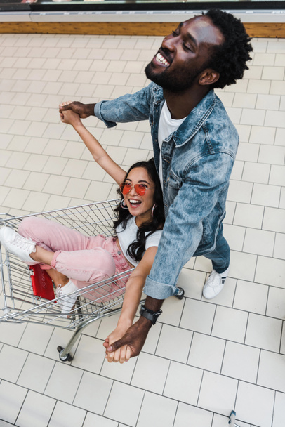 bovenaanzicht van gelukkig Aziatisch meisje in zonnebril zitten in winkelwagen en handen vasthouden met knappe Afro-Amerikaanse man  - Foto, afbeelding