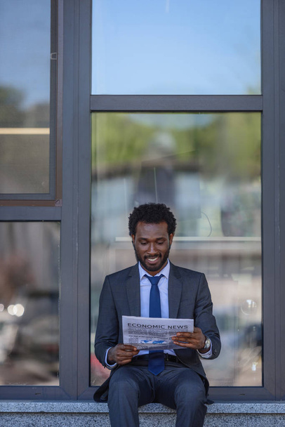 sonriente hombre de negocios afroamericano leyendo el periódico mientras está sentado cerca del edificio de oficinas con fachada de vidrio
 - Foto, imagen