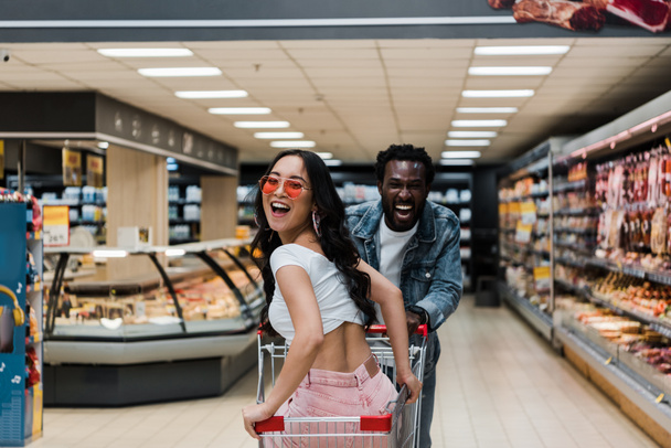 Gelukkig Aziatische meisje in zonnebrillen kijken naar camera terwijl zitten in winkelwagen in de buurt van positieve Afro-Amerikaanse man  - Foto, afbeelding
