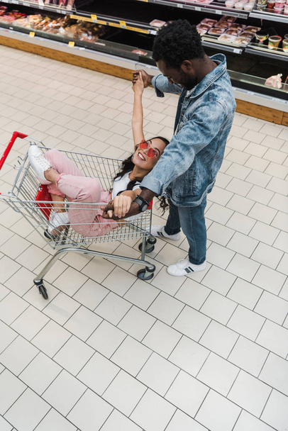 overhead view of positive asian girl in sunglasses sitting in shopping cart and holding hands with african american man  - Фото, зображення
