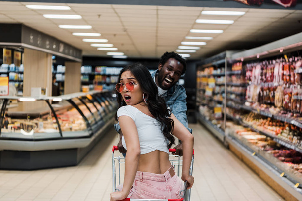 verrast Aziatische meisje in zonnebril zitten in winkelwagen in de buurt van positieve Afro-Amerikaanse man  - Foto, afbeelding