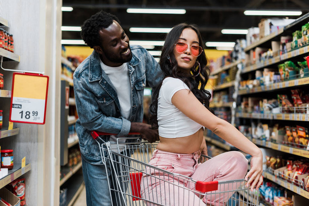 handsome african american man looking at stylish asian woman in sunglasses sitting in shopping cart - Zdjęcie, obraz