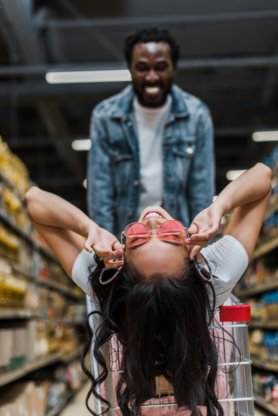 selective focus of cheerful asian woman touching sunglasses and sitting in shopping cart near happy african american man  - Foto, Bild