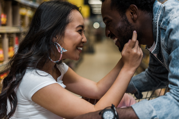 selective focus of happy asian girl touching face of handsome and cheerful african american man  - Φωτογραφία, εικόνα