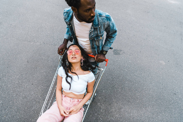 overhead view of african american man standing near asian girl sitting in shopping trolley outside  - Fotografie, Obrázek