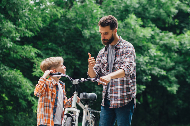 Knappe baard vader praat met zijn zoon fietsen en toont duim omhoog in zomerpark groene bladeren op achtergrond. - Foto, afbeelding