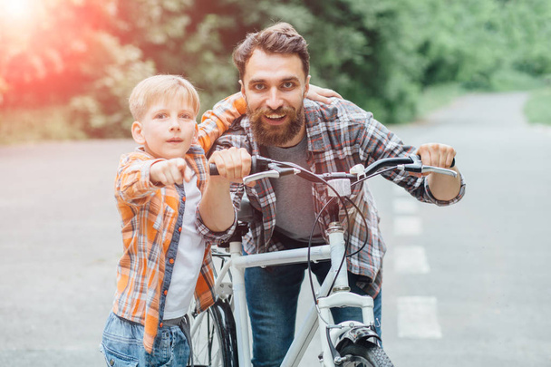 Πατέρας και γιος διασκεδάζουν μαζί ιππασία ποδήλατο μαζί, περπατώντας σε raod σε καταπράσινο πάρκο ή δάσος. Το παιδί δείχνει με τα χέρια μπροστά, και τα δύο χαμόγελα. Αίσθηση ψυχαγωγίας περιπέτειας. - Φωτογραφία, εικόνα