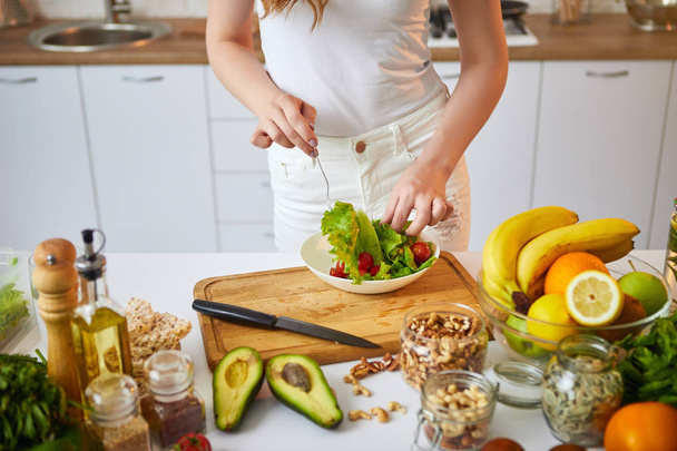 Jonge gelukkige vrouw bereidt smakelijke salade in de prachtige keuken met groene verse ingrediënten binnenshuis. Gezond eten en dieet concept. Loosing gewicht - Foto, afbeelding