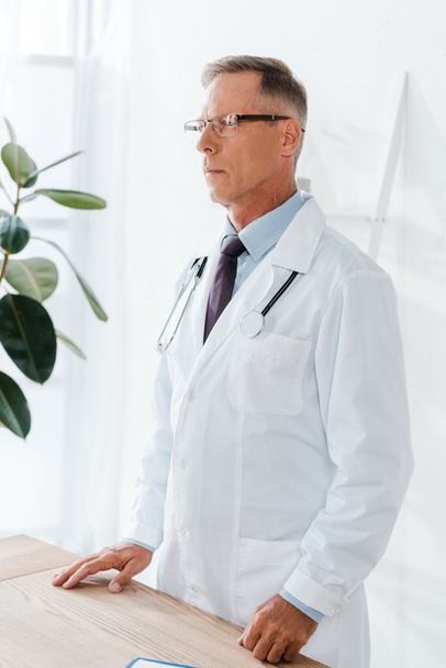 όμορφος γιατρός με λευκό παλτό και γυαλιά στέκεται κοντά στο γραφείο  - Φωτογραφία, εικόνα