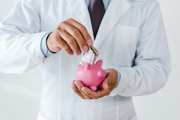 cropped view of man in white coat putting dollar banknote in piggy bank on white - Foto, Imagen