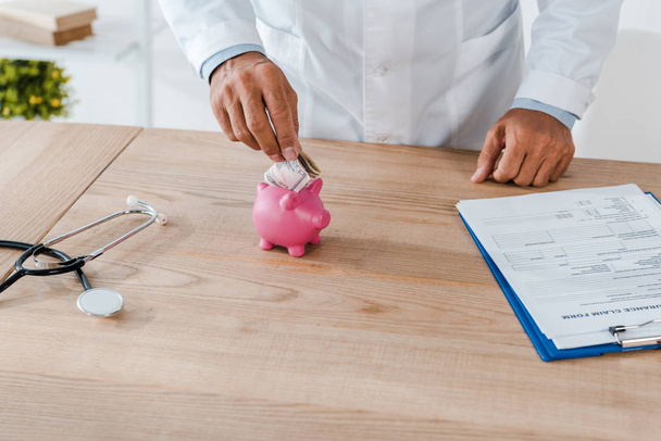 cropped view of man in white coat putting dollar banknote in piggy bank near clipboard and stethoscope  - Fotografie, Obrázek