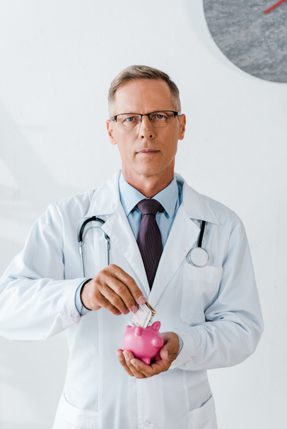 man in white coat putting dollar banknote while holding piggy bank - Photo, Image