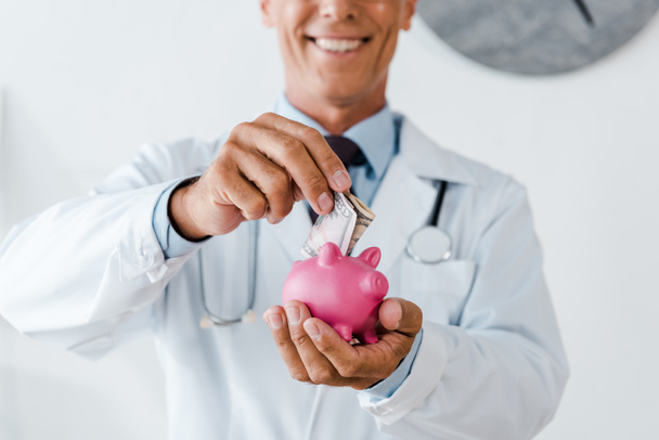 cropped view of cheerful man in white coat putting dollar banknote while holding piggy bank - Fotoğraf, Görsel