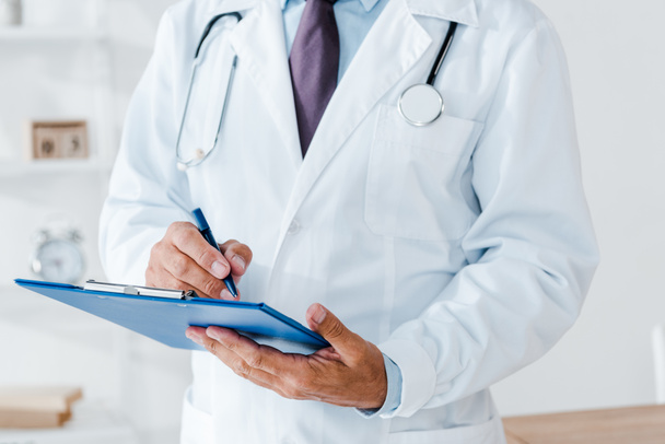cropped view of doctor in white coat holding clipboard and writing diagnosis  - Fotografie, Obrázek