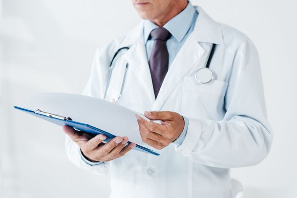 Ausgeschnittene Ansicht eines Arztes in weißem Kittel mit Seite und Klemmbrett in der Klinik  - Foto, Bild