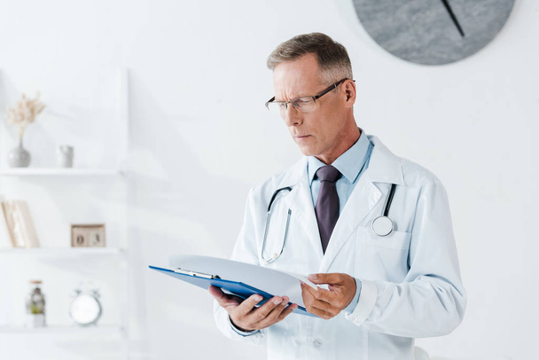 handsome man in white coat looking at clipboard in clinic  - Foto, afbeelding