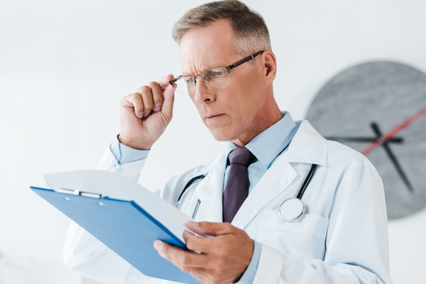 επιλεκτική εστίαση του όμορφου γιατρού σε λευκό παλτό κοιτάζοντας το Πρόχειρο και αγγίζοντας τα γυαλιά στο νοσοκομείο  - Φωτογραφία, εικόνα