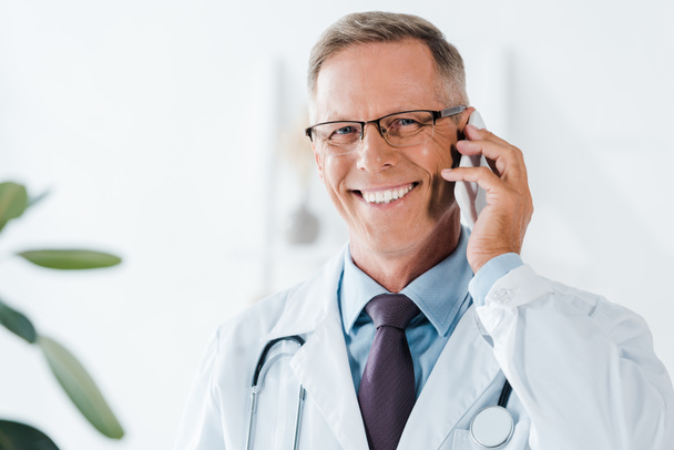gelukkige dokter kijken naar camera en praten op smartphone in kliniek  - Foto, afbeelding