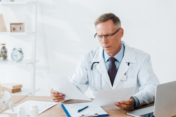 médico bonito em óculos olhando para documentos na clínica
  - Foto, Imagem