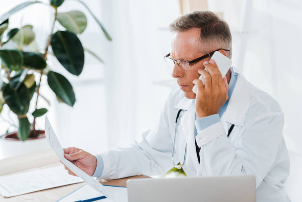 doctor en gafas y bata blanca hablando en smartphone y mirando el documento
  - Foto, imagen