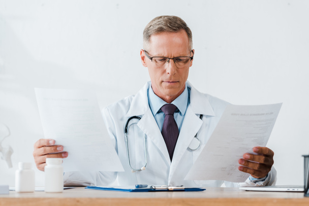 doctor en gafas y bata blanca con estetoscopio mirando documentos
  - Foto, imagen
