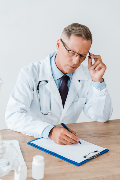 doctor in white coat touching glasses and writing diagnosis in clinic  - Foto, afbeelding