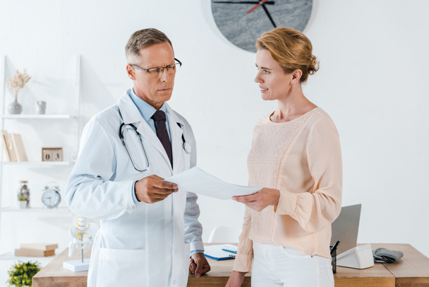 doctor in glasses and white coat looking at document near attractive woman  - Foto, imagen