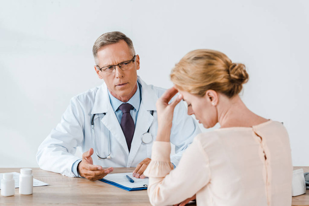 selective focus of doctor in glasses looking at upset woman touching head while sitting near table  - Foto, Imagen