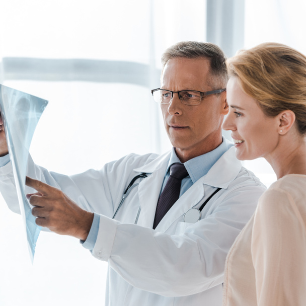 przystojny lekarz w okulary wskazując blisko szczęśliwy kobieta patrząc na x-ray  - Zdjęcie, obraz
