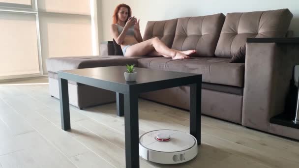 Smart House. Reinigung der Häuser Staubsaugerroboter, wenn Frauen auf Sofas Mobiltelefone spielen - Filmmaterial, Video