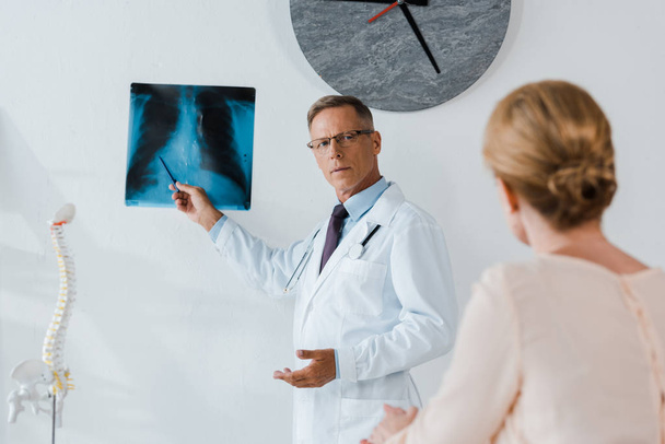 selektivní zaměření lékaře v bílém plášti a skleničkách přidržel pero u rentgenové a ženy na klinice  - Fotografie, Obrázek