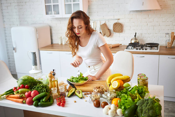 Young happy woman eating salad in the beautiful kitchen with green fresh ingredients indoors. Healthy food concept - Фото, изображение