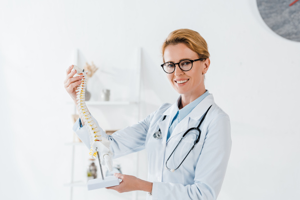cheerful doctor in glasses holding spine model and smiling in hospital  - 写真・画像