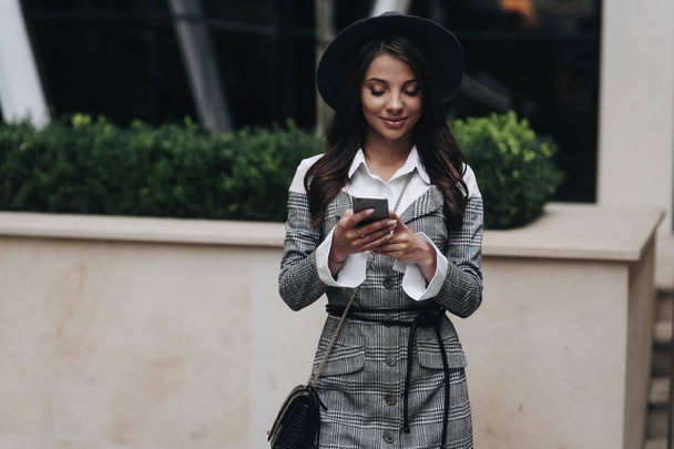 Šťastná žena nosí v šedém kabátě a klobouku pomocí inteligentního telefonu v s - Fotografie, Obrázek