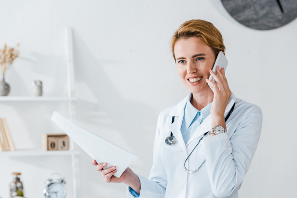 médico alegre sosteniendo papel y sonriendo mientras habla en el teléfono inteligente
  - Foto, Imagen