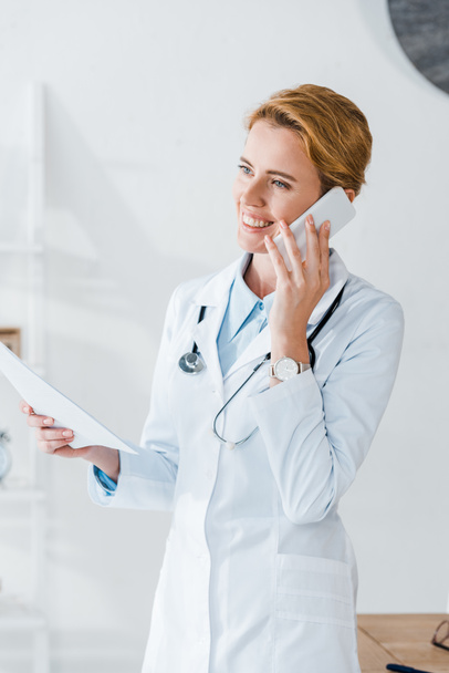 happy doctor holding paper and smiling while talking on smartphone  - Foto, Bild