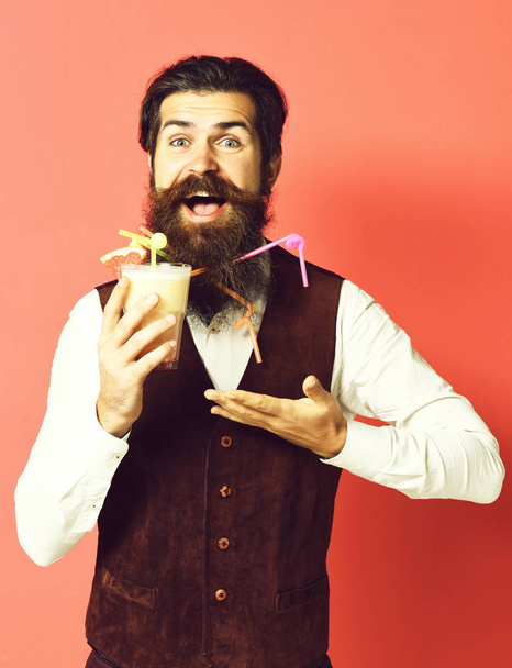handsome bearded man with long beard and mustache has stylish hair on smiling face holding glass of alcoholic beverage in vintage suede leather waistcoat on red studio background - Фото, зображення