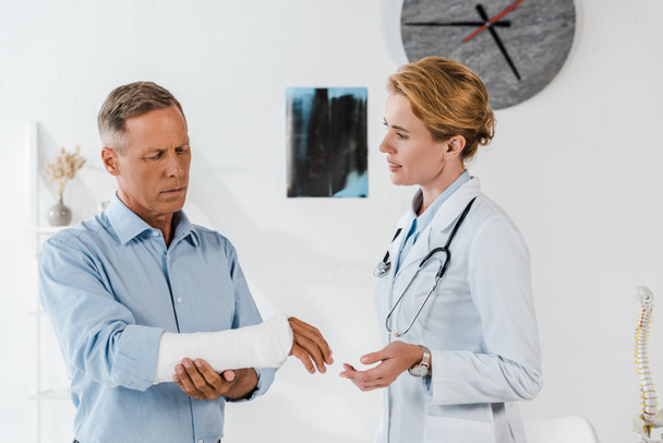 attractive doctor looking at handsome man with broken arm  - Photo, Image