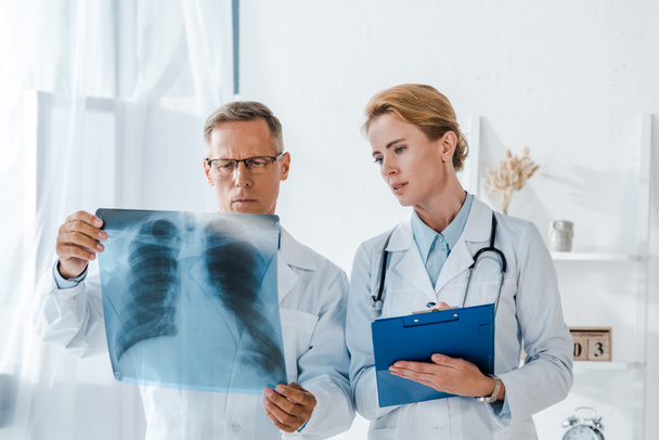 atractivo médico sujetando portapapeles y mirando rayos X cerca de compañero de trabajo
  - Foto, Imagen